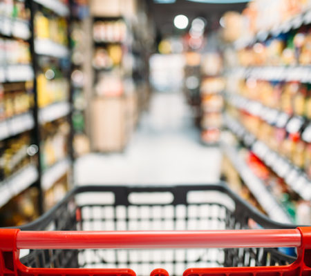 Shopping cart between shelves in food store, nobody. Rows of products in supermarket